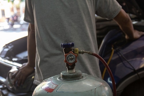 DIY A/C Recharging vs. Professional A/C Repair | Accelerate Auto Repair in Rowlett, TX. Closeup image of a tank valve of a freon bottle with auto mechanic working on a car air conditioner in the background.