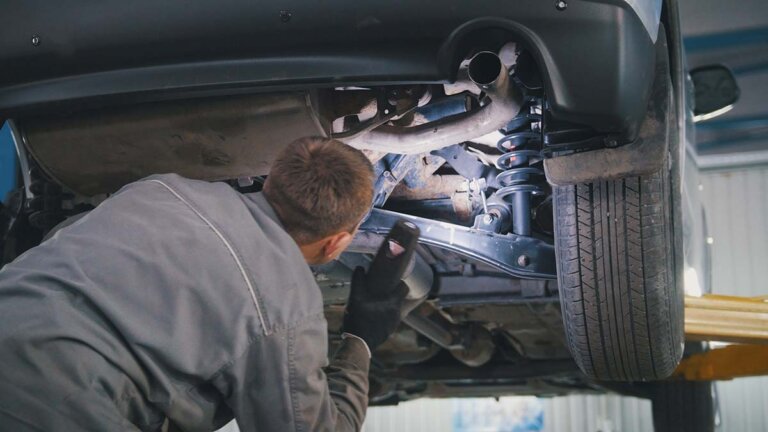 Person inspecting a vehicle’s front suspension and control arm area on a lift.