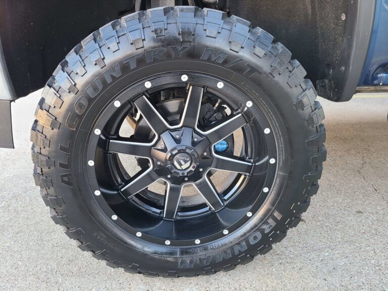Close-up of a black aftermarket wheel and off-road tire mounted on a truck, with the wheel and hub assembly visible.