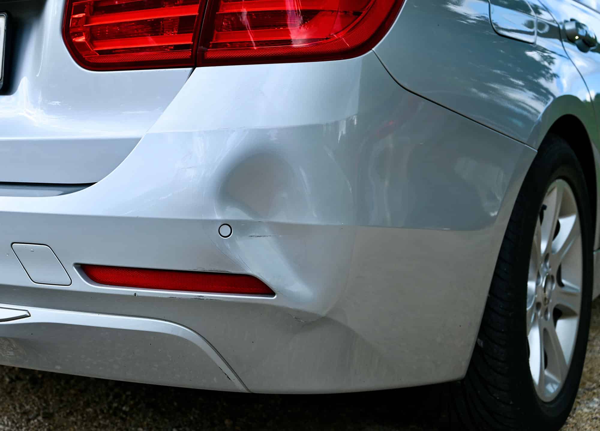 Dented and scraped rear bumper on a silver sedan, viewed from the side.